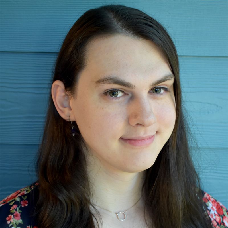 A photograph of Tara Wilkins, a woman with long brown hair wearing a necklace and earrings, standing in front of a blue wall.