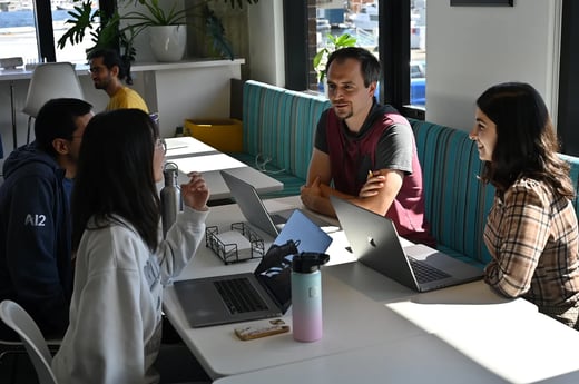A photograph of four people sitting together at a cafeteria table, with laptops in front of them.